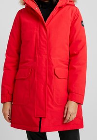 Rode parka met een bontgevoerde capuchon, twee voorzakken en een volledige ritssluiting. Gemaakt van duurzaam materiaal met verstelbare manchetten.