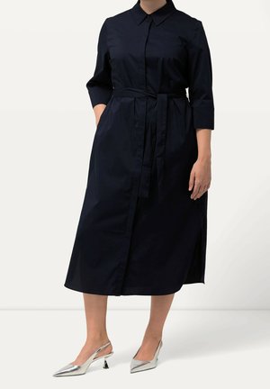 Femme portant une robe chemise midi noire à manches trois-quarts, ceinture nouée à la taille, et des escarpins argentés à bout pointu avec bride arrière.