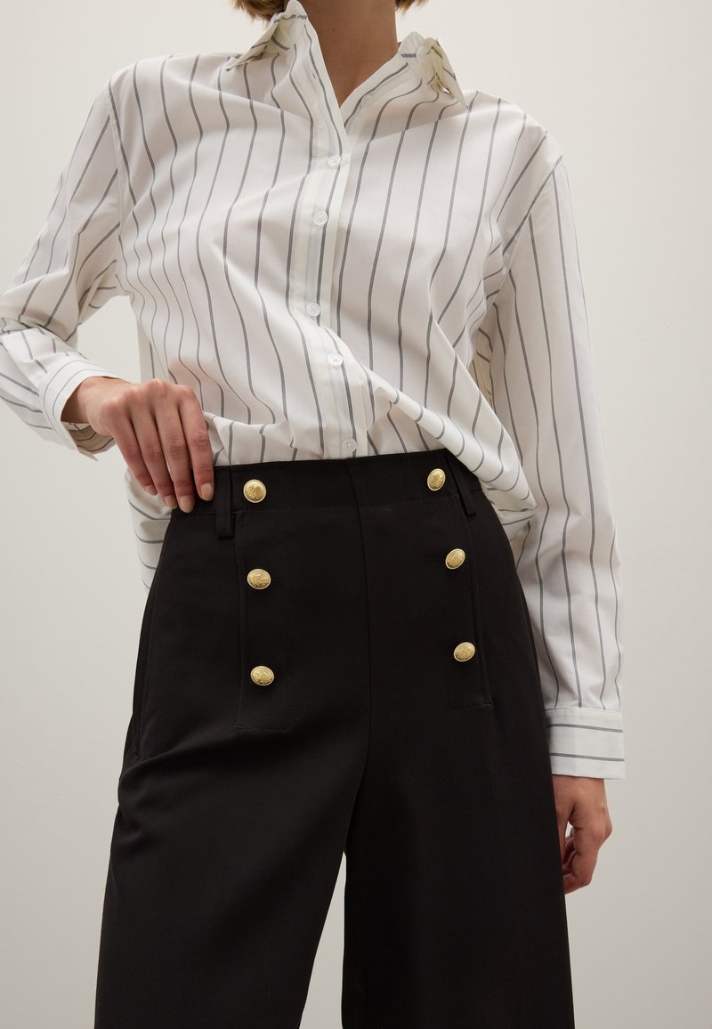 Witte gestreepte overhemd met knopen en een kraag, gecombineerd met hoge zwarte broeken met gouden knopen en een getailleerd ontwerp.