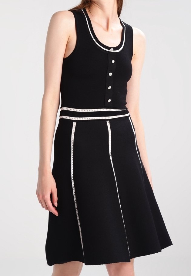 Femme portant une robe noire sans manches avec un liseré blanc, des boutons décoratifs sur le corsage et des rayures blanches verticales sur la jupe évasée.