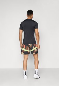 Hombre de pie de espaldas, vistiendo una camiseta negra ajustada, pantalones cortos abstractos multicolores con rayas amarillas, calcetines blancos y zapatillas blancas.