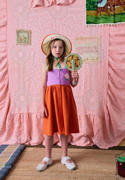 Fille en robe violette et orange portant un chapeau de paille, tenant un éventail tressé, debout devant un fond en tissu rose à motifs.
