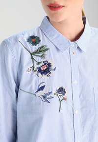 Camicia button-up a righe blu con ricami floreali di diversi colori sulla spalla sinistra e sul petto, dotata di colletto classico e tasca.