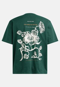 Grünem Baumwoll-T-Shirt mit einem weißen Blumenmuster auf der Rückseite, mit Text darüber und darunter in Orange und Weiß. Kurze Ärmel und lässige Passform.