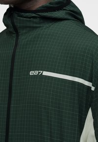 Σκούρο πράσινο μπουφάν με κουκούλα και φωτεινό σχέδιο πλέγματος, μαύρο φερμουάρ, λευκή οριζόντια ρίγα και λογότυπο "ea7" στο στήθος.