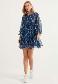 Blauwe bloemenjurk van doorschijnende stof met lange mouwen en een elastische tailleband, met een gelaagde rok, gecombineerd met witte sneakers.