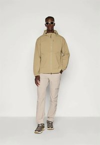 Μπεζ αδιάβροχο μπουφάν με μπροστινό φερμουάρ, μακριά μανίκια και ρυθμιζόμενη κουκούλα. Συνδυάζεται με ανοιχτό μπεζ παντελόνι και σκούρα υποδήματα.