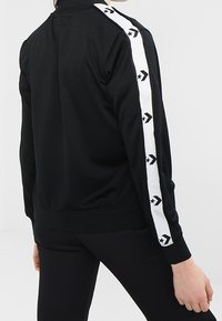 Black track jacket with a white stripe featuring black arrows and stars along the sleeves, made from a smooth, stretchy fabric.