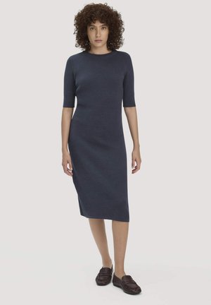 Frau mit lockigem Haar, die ein figurbetontes, knielanges Kleid in Marineblau und braune Loafers trägt, steht vor einem schlichten, hellen Hintergrund.