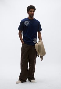 Μπλούζα μπλε ναυτικού βαμβακερή με λευκά μοτίβα ήλιου· συνδυάζεται με καφέ παντελόνια φαρδιάς γραμμής και κρατάει ένα μπεζ πουλόβερ. Χαλαρή εφαρμογή.