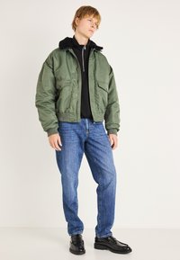 Chaqueta bomber verde con cuello de piel negra, usada sobre una camiseta negra, combinada con jeans azules y zapatos negros. Cuenta con bolsillos con cremallera.
