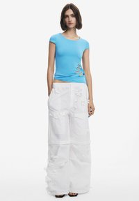 Haut ajusté bleu à manches courtes, avec une découpe sur le côté ; associé à un pantalon blanc à jambes larges orné d'appliqués floraux texturés.