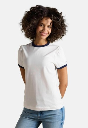 Young woman with curly hair smiling, wearing white short-sleeve T-shirt with navy trim and light blue jeans, standing against white background.