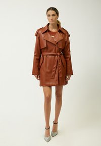 Bruine faux leren trenchcoat met een ceintuur in de taille, reverskraag en schouderepauletten. Gekozen over een rode trui. Sprankelende hakken maken de look compleet.