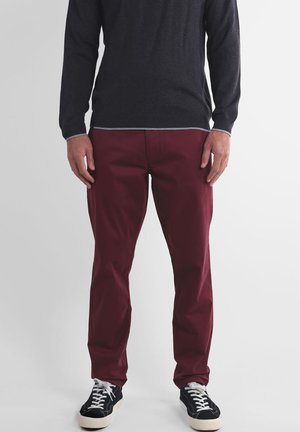 Pantalon bordeaux en coton, avec une coupe droite et des poches latérales. Porté avec un pull foncé et des baskets noires.
