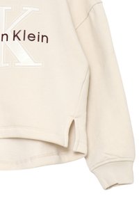 Felpa beige con vestibilità rilassata, caratterizzata da una grande "K" bianca e il testo "Calvin Klein" in marrone scuro. Maniche a costine e spacchetti laterali all'orlo.