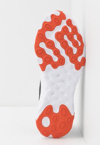 La semelle de la chaussure présente une base blanche avec des accents en caoutchouc rouge et des motifs circulaires. Texture flexible, conçue pour l'adhérence et la durabilité.