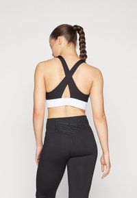 Črna športna modrček z križnim dizajnom na hrbtu in belim pasom; v kombinaciji z visokim pasom črnimi leggins, ki imajo teksturirane mrežaste podrobnosti.