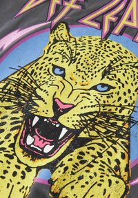 Grafična majica s slikom divjega rumenega leva z črnimi madeži in modrimi očmi. Ozadje ima roza in modre poudarke z drznim besedilom.