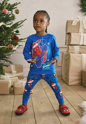 Barn i blå Spider-Man-nattøj og røde hjemmesko står ved juletræet og indpakkede gaver i et festligt rum.