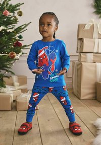 Bambino in pigiama blu di Spider-Man e pantofole rosse sta accanto all'albero di Natale e ai regali incartati in una stanza festosa.
