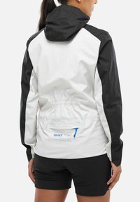 Person in einer schwarz-weißen Kapuzenjacke mit einem Reißverschluss-Rückentasche und schwarzen Shorts, die von der Kamera abgewandt ist.