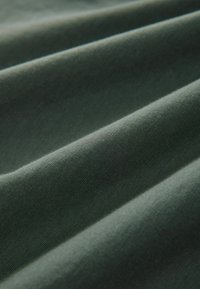 Tmavě zelená látka, hladký povrch, mírně elastická, s viditelnými záhyby, které vytvářejí přirozené sklady. Žádné vzory ani kovové prvky nejsou přítomny.