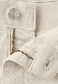 Tissu en denim beige avec une fermeture éclair métallique partiellement ouverte et un bouton argenté portant la mention « MNG DENIM ».