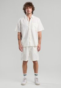 Camicia bianca in lino a maniche corte con taschino, abbinata a pantaloni corti coordinati. Indossata con sneakers bianche e calzini alla caviglia a righe.