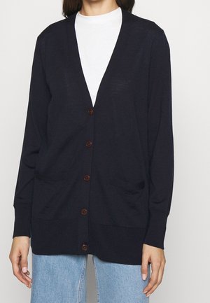 Femme portant un cardigan bleu marine boutonné par-dessus un top blanc et un jean bleu clair, debout devant un fond blanc uni.