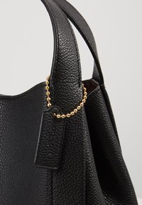 Sac à main en cuir noir grainé avec de larges anses et une petite étiquette rectangulaire en cuir attachée par une chaîne à billes dorée.