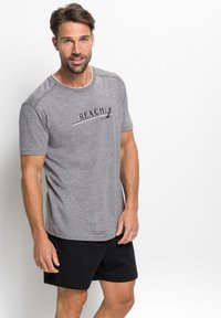Graues Baumwoll-T-Shirt mit kurzen Ärmeln, mit einem schwarzen grafischen Text "REACH" über der Brust. Kombiniert mit schwarzen Shorts.