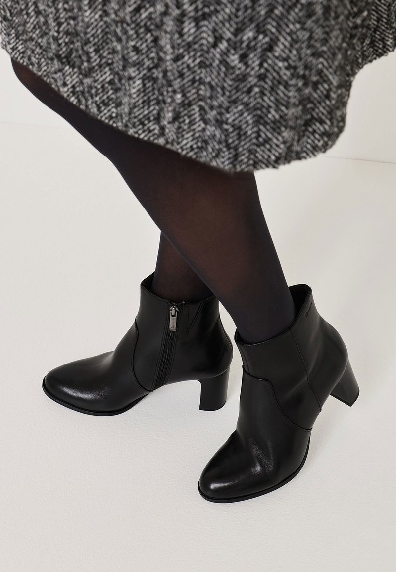 Bottines en cuir noir avec un bout pointu, une fermeture éclair sur le côté et un talon épais, présentées sur un fond clair.
