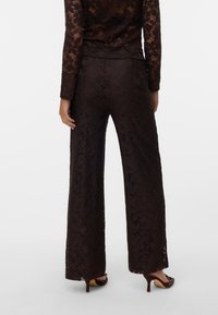 Femme portant un pantalon en dentelle noire à motifs floraux et un haut à manches longues assorti, vue de dos, debout sur un fond blanc uni.