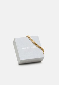 Aukso apyrankė gulėja ant tekstūruoto pilko dėžutės, kurioje baltuoju spaudu užrašyta "MICHAEL KORS". Dėžutė turi švelnų, matinį paviršių.