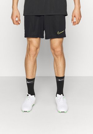 Mandeben iført sorte Nike-shorts og sorte Nike-strømper med hvide Nike-fodboldstøvler på en ensfarvet grå gulv og hvid baggrund.