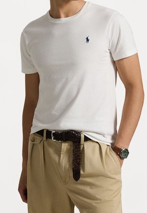 Mand iført en simpel hvid kortærmet T-shirt med lille marineblåt logo, beige bukser med vævet brunt bælte og ur med mørkegrøn urskive.