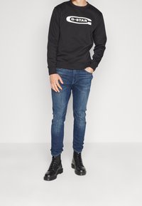 Muž ve černé mikině G-STAR, modrých úzkých džínách s šitím na kolenou a černých vojenských kotníkových botách se šněrováním, stojící na bílém pozadí.