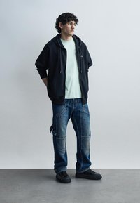 Έφηβος άντρας όρθιος, φοράει μαύρο φούτερ με φερμουάρ, ανοιχτόχρωμο μπλουζάκι μέντας, φθαρμένα σκούρα μπλε τζιν με μπαλώματα και μαύρα αθλητικά παπούτσια.