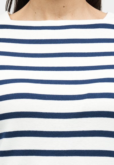 Ριγέ μπλουζάκι με οριζόντιες ναυτικές και λευκές ρίγες, κατασκευασμένο από μαλακό βαμβάκι. Διαθέτει λαιμόκοψη τύπου σκάφος και ριμπ υφή.