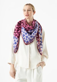 Écharpe en forme de triangle avec des motifs de cœurs roses et bleus, ornée d'une bordure rouge contrastante, fabriquée en tissu doux. Portée lâchement sur une chemise blanche.