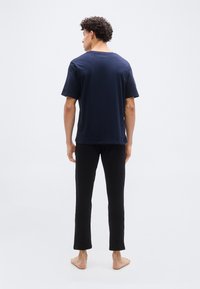 Άνδρας που στέκεται ξυπόλυτος, με την πλάτη γυρισμένη, φορώντας μπλε σκούρο μπλουζάκι και μαύρο παντελόνι, σε απλό λευκό φόντο.