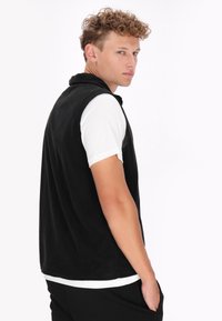 Schwarzige Fleece-Weste mit hohem Kragen und Reißverschluss, mit Seitentaschen, getragen über einem weißen Hemd, vor einem schlichten weißen Hintergrund präsentiert.
