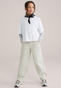 Ung person står med hænderne i lommerne, iført hvid oversized T-shirt, grå langærmet trøje under, beige bukser, sneakers og sort halstørklæde.