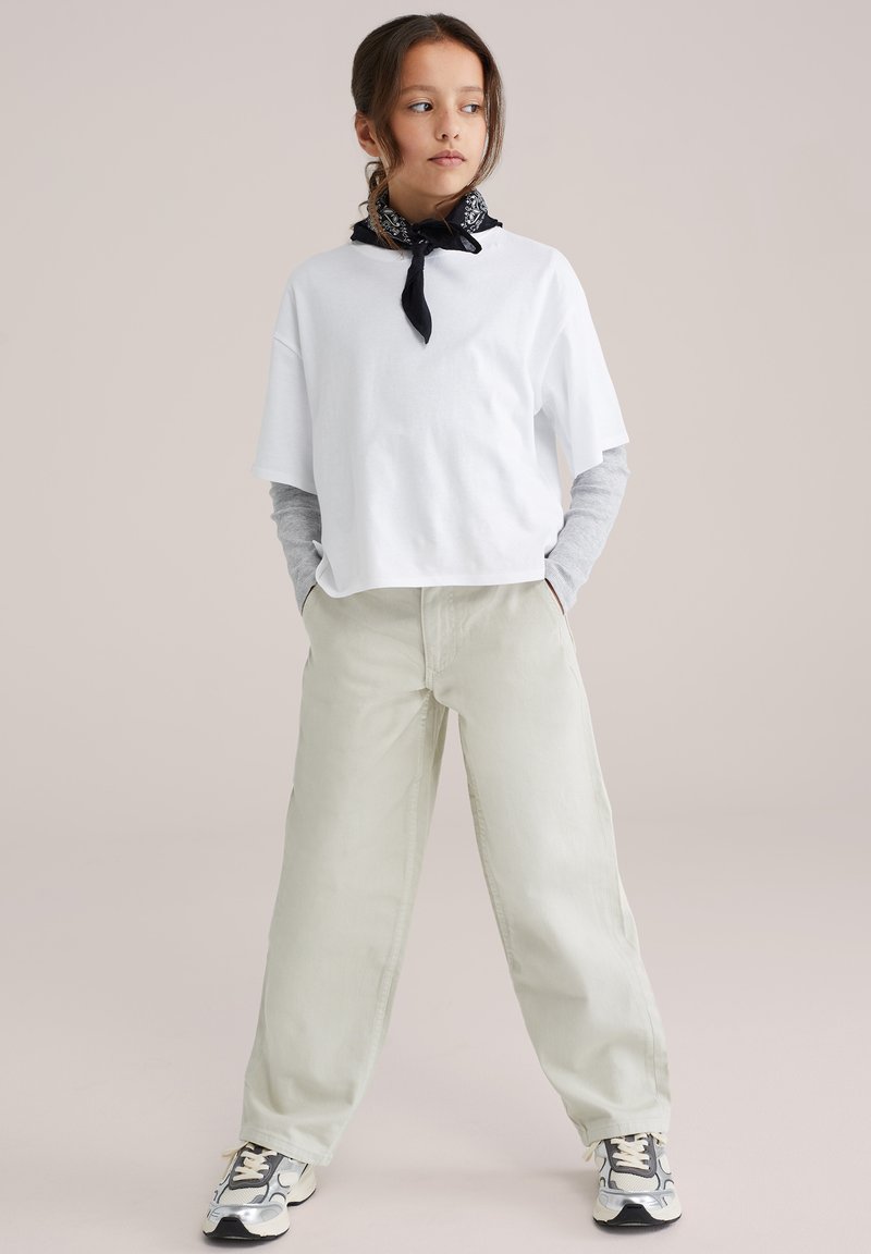 Ung person står med hænderne i lommerne, iført hvid oversized T-shirt, grå langærmet trøje under, beige bukser, sneakers og sort halstørklæde.