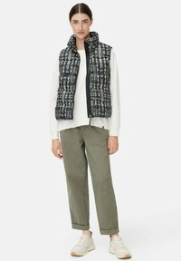 Vrouw in een zwart-wit geblokte pufferjack over een wit langemouwen shirt, olijfgroene broek en witte sneakers, staand.