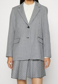 Blazer de cuadros en gris y blanco con solapa de muesca, cierre de un solo botón y dos bolsillos frontales. Combinado con una falda plisada a juego.