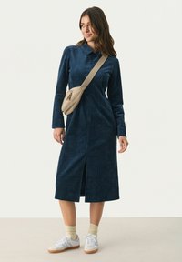 Dunkelblaues Corduroykleid mit langen Ärmeln, einem Kragen und einem Schlitz vorne. Kombiniert mit einer beigen Umhängetasche und weißen Sneakern.