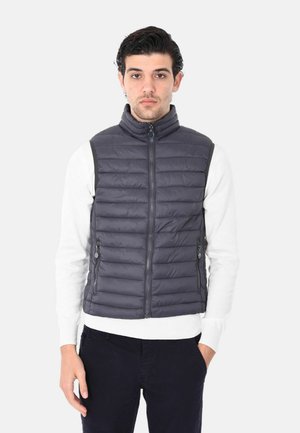 Gilet imbottito grigio con colletto alto, zip frontale e tasche laterali. Indossato sopra un maglione bianco e abbinato a pantaloni scuri. Texture liscia.