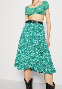 Vihreä kukallinen crop-top, jossa on neliökaula-aukko, yhdistettynä yhteensopivaan hameeseen, jossa on kerrostettu helma ja musta vyö hopeasolmulla.